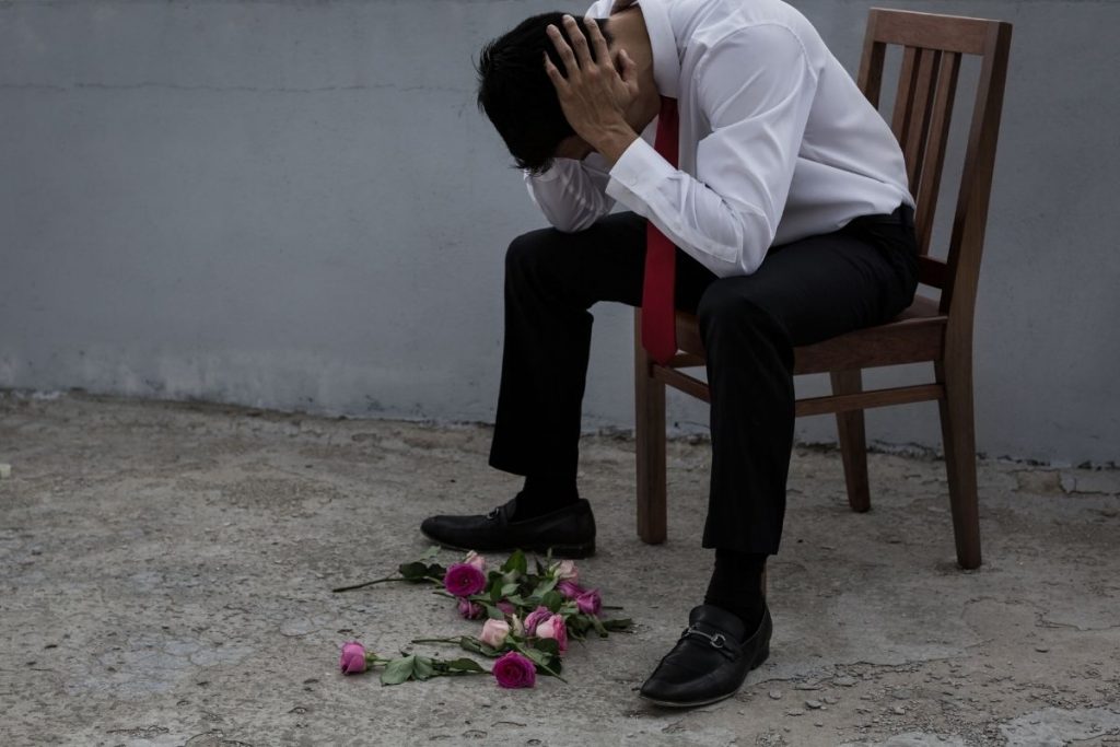 Man sitting in a chair holding his head with flowers strewn at his feet.